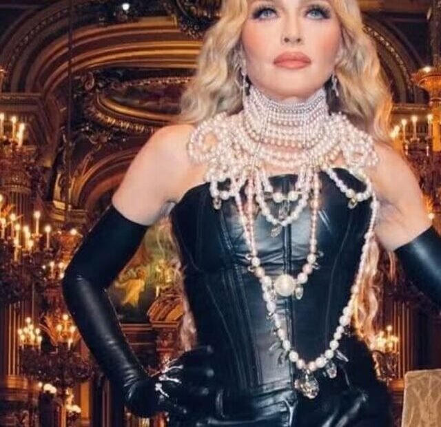 Madonna posing in an opulent room, dressed in a black leather dress and adorned with multiple pearl necklaces and heavy makeup, exuding glamour.