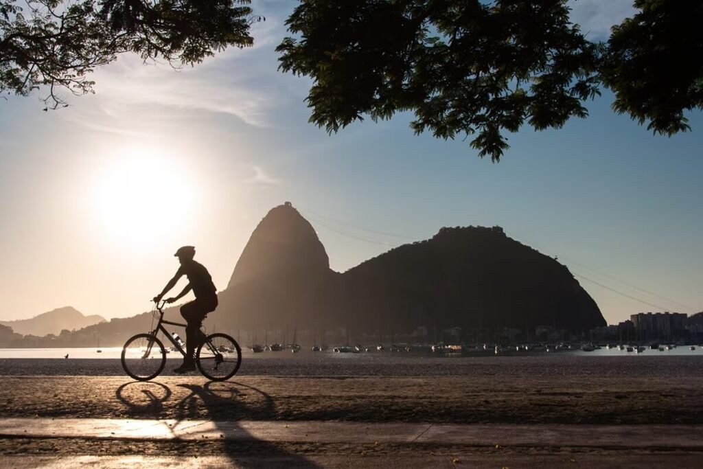 A cyclist enjoying a scenic ride along the beachfront promenade in Rio de Janeiro, with Sugarloaf Mountain in the background and the golden sun setting over the city skyline.